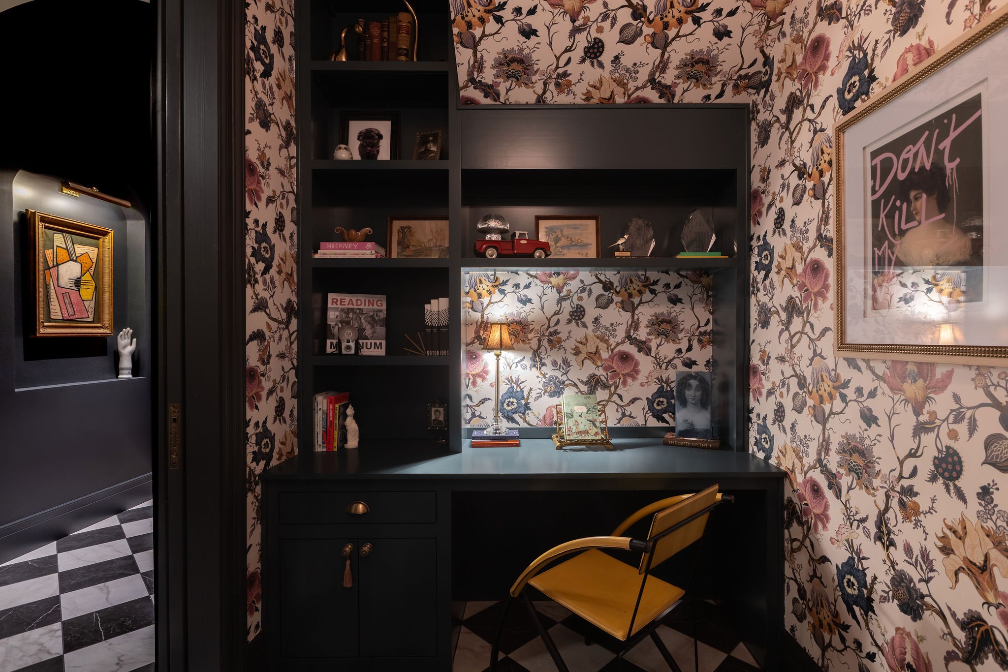 Office at night with floral wallpaper, navy built-ins, and view to a lit painting in the hall