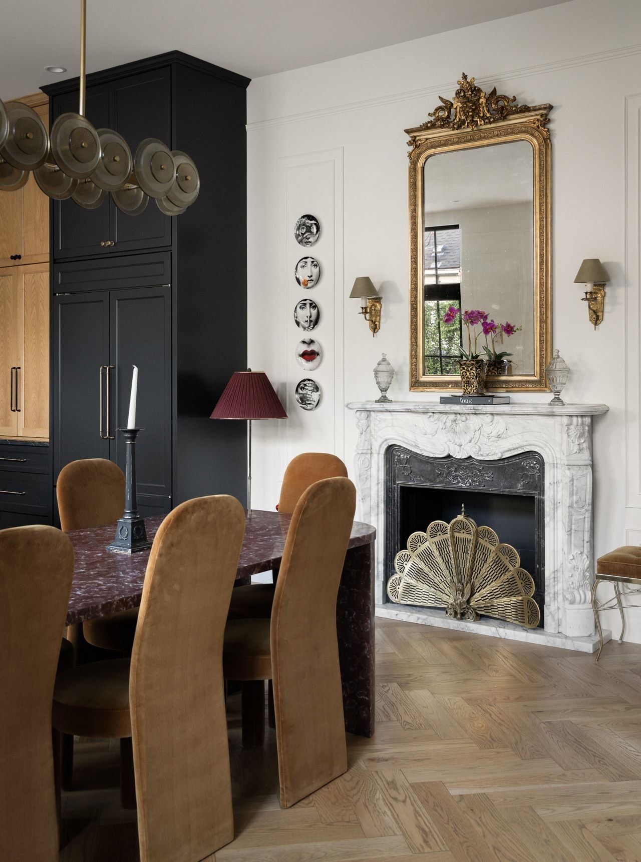 Marble fireplace with a gilded mirror and wall plates, wood cabinetry, and caramel dining chairs on herringbone floors
