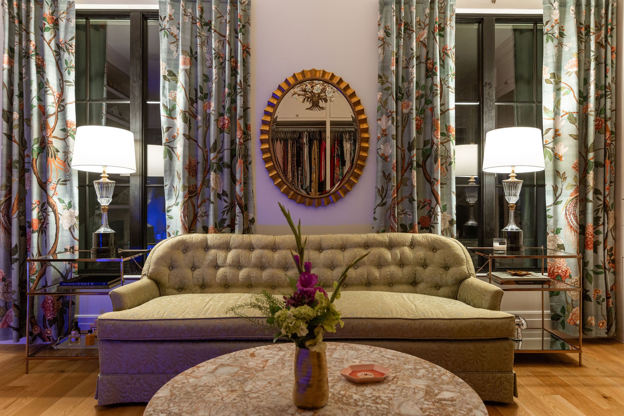 Green tufted sofa between floral curtains and glowing lamps with a round rattan mirror